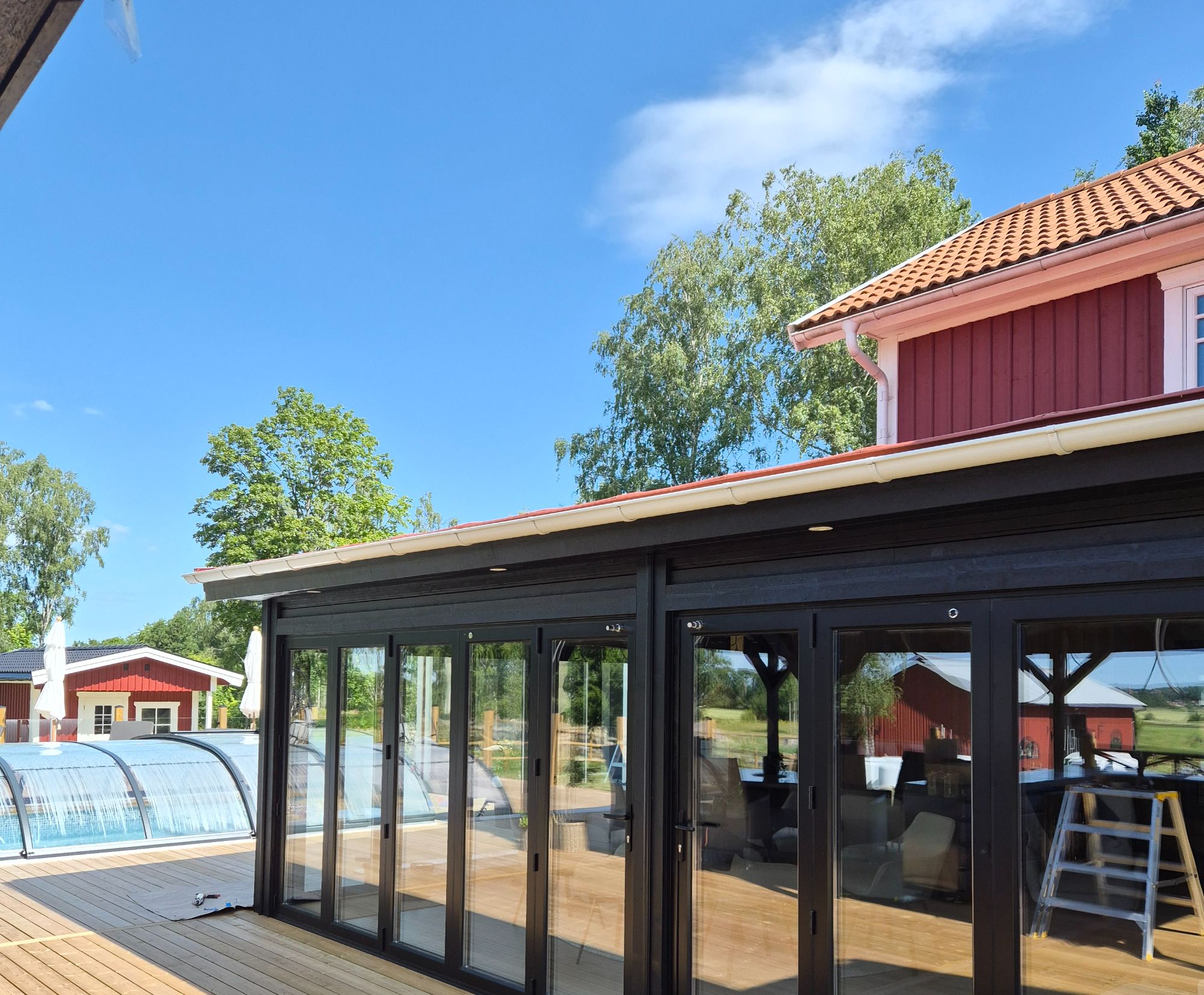 Vikdörrspartier i glas under svartmålad takfot, med trädäck och pooltak intill