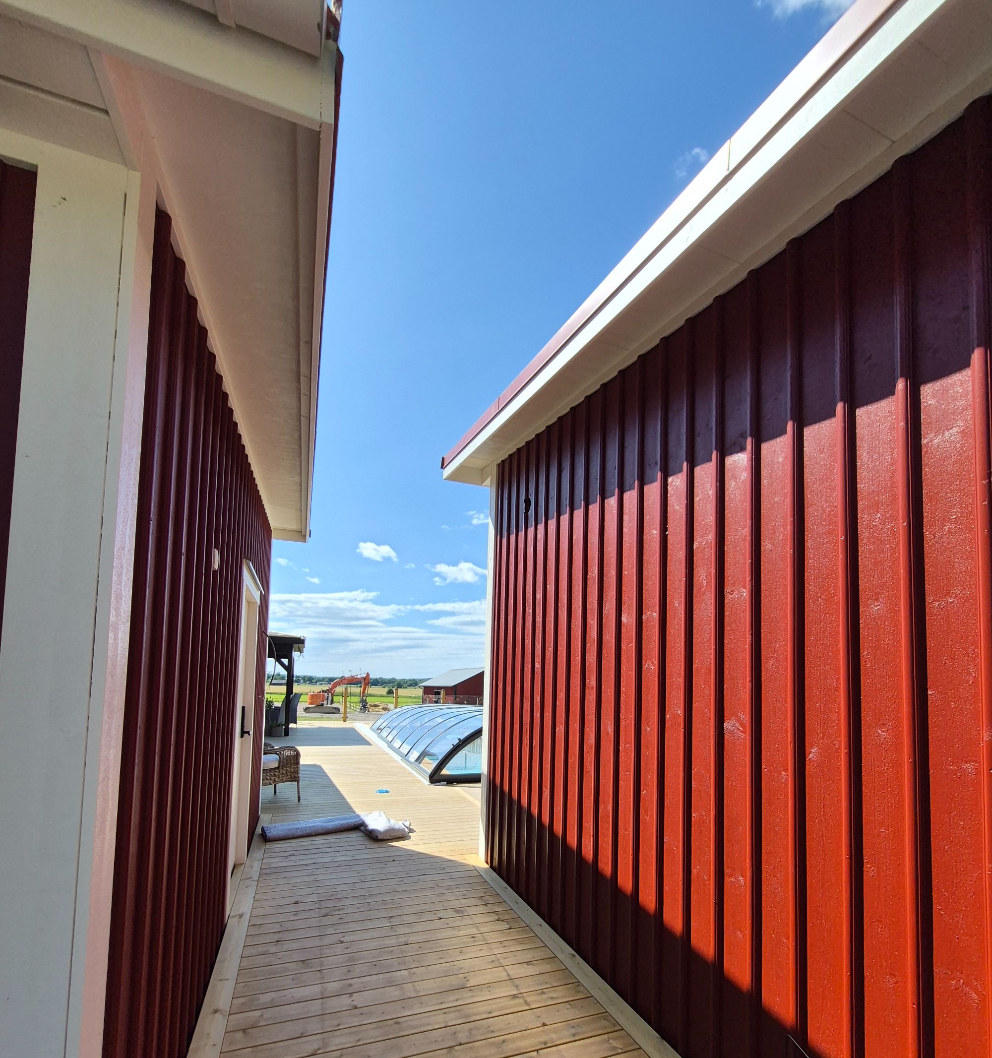 Trädäck mellan två rödmålade träfasader, pooltaket skymtar längre fram